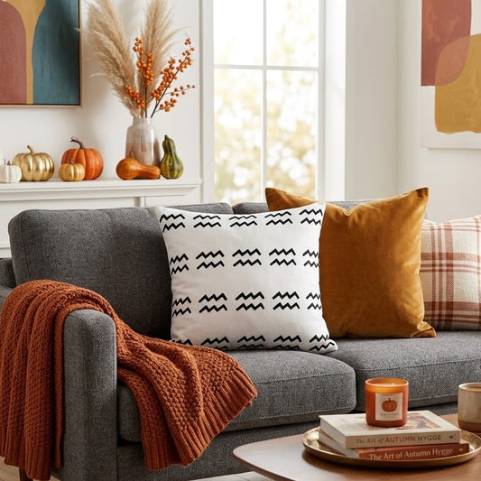 A gray upholstered armchair in a styled living room, with an orange knit throw draped over one arm, a white pillow with a black aquarius pattern, a mustard yellow pillow, and a plaid accent pillow. A wooden coffee table, books, a candle, and a mug sit in front of the chair.