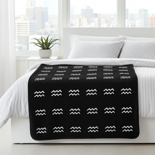 Black blanket with white Aquarius patterns draped over a bed in a bright room with cityscape view.