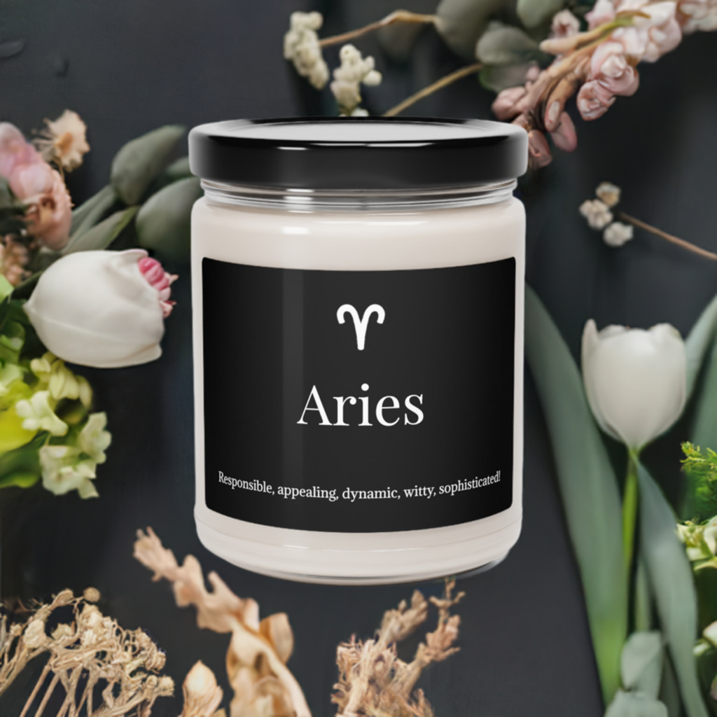 A white scented soy candle with a black label that reads 'Aries' placed in a glass jar with a black lid, surrounded by flowers in the background.