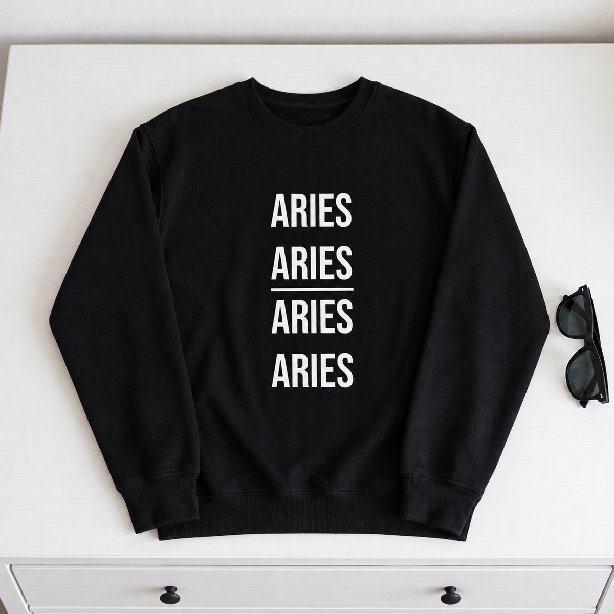 A black crewneck sweatshirt with 'ARIES ARIES ARIES ARIES' printed in white block letters on the front, laid flat on a white surface next to a pair of sunglasses.