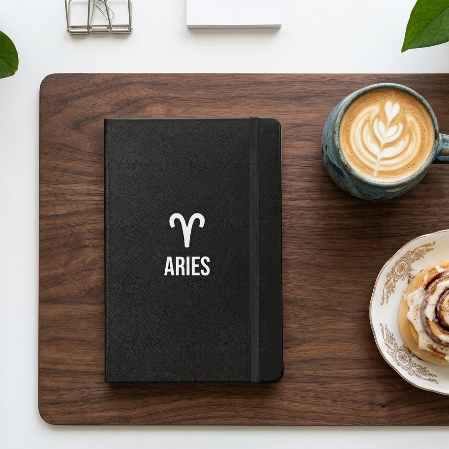 A black hardcover notebook with a white Aries zodiac symbol and the word 'ARIES' on the cover, placed on a dark wood tray. The tray is set on a white desk alongside a cup of latte art coffee, a pastry on a saucer, and green leaves.
