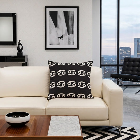 A black throw pillow with a white astrological symbol pattern, styled on a cream leather sofa in a modern living room setting.