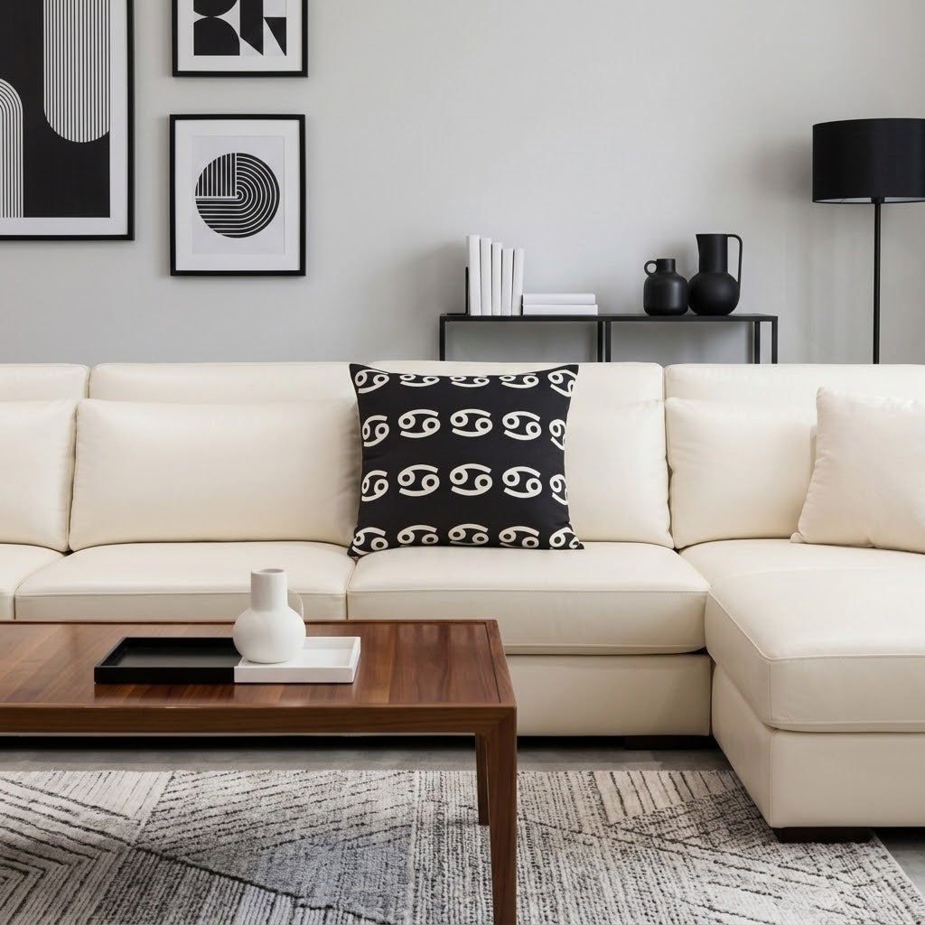 A modern living room scene featuring a cream-colored sectional sofa with a black throw pillow printed with a white interlocking logo pattern, set in front of a dark wood coffee table and a gray patterned rug.