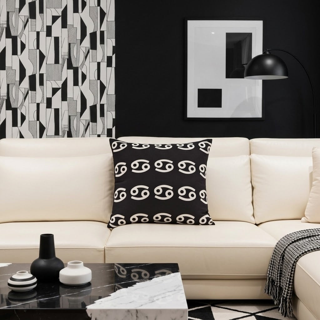 A cream-colored sectional sofa in a modern living room setting, featuring a black throw pillow with a white astrological symbol pattern. The room has a black accent wall with abstract art, a black floor lamp, and a marble coffee table in the foreground.
