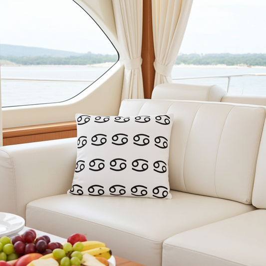 A square white throw pillow with a black astrological Cancerian zodiac symbol pattern, resting on a cream-colored yacht sofa with a fruit platter in the foreground and water scenery in the background.