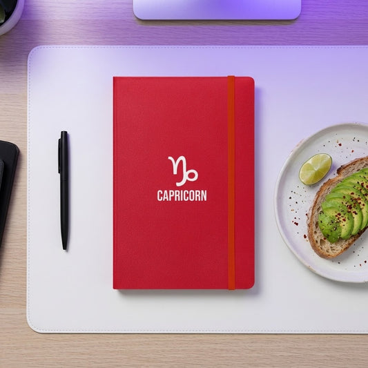 A red hardcover notebook with a white Capricorn zodiac symbol and text on the front, placed on a white desk mat over a light wood desk. A black pen is to the left, and a plate with avocado toast and a lime wedge is to the right.