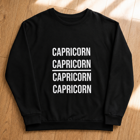 A black crewneck sweatshirt with 'CAPRICORN' printed in white block letters four times on the front, laid flat on a light brown wooden floor.