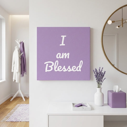 A square lavender wall art print with the phrase 'I am Blessed' in white script, displayed on a white wall in a styled room with a vanity, mirror, and lavender accessories.