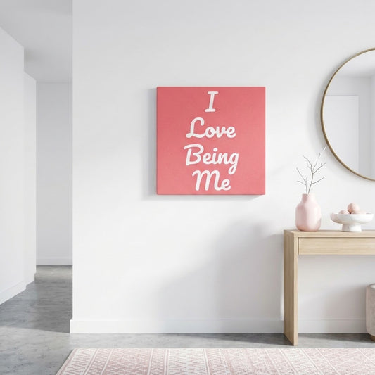 A square canvas wall art print with the text 'I Love Being Me' in white script on a coral pink background, displayed on a white wall in a modern, minimalist room with a light wood console table and a round mirror.