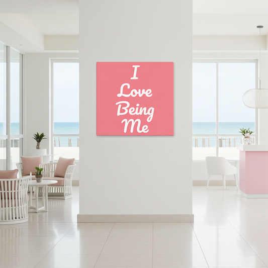 A square pink wall art print with white text reading 'I Love Being Me' is displayed on a white wall in a bright, modern interior with an ocean view in the background.