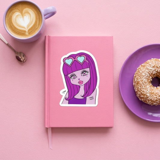 A pink hardcover notebook with a die-cut sticker of a cartoon girl with purple hair and heart-shaped glasses, styled on a pink surface with a coffee cup, spoon, and donut.