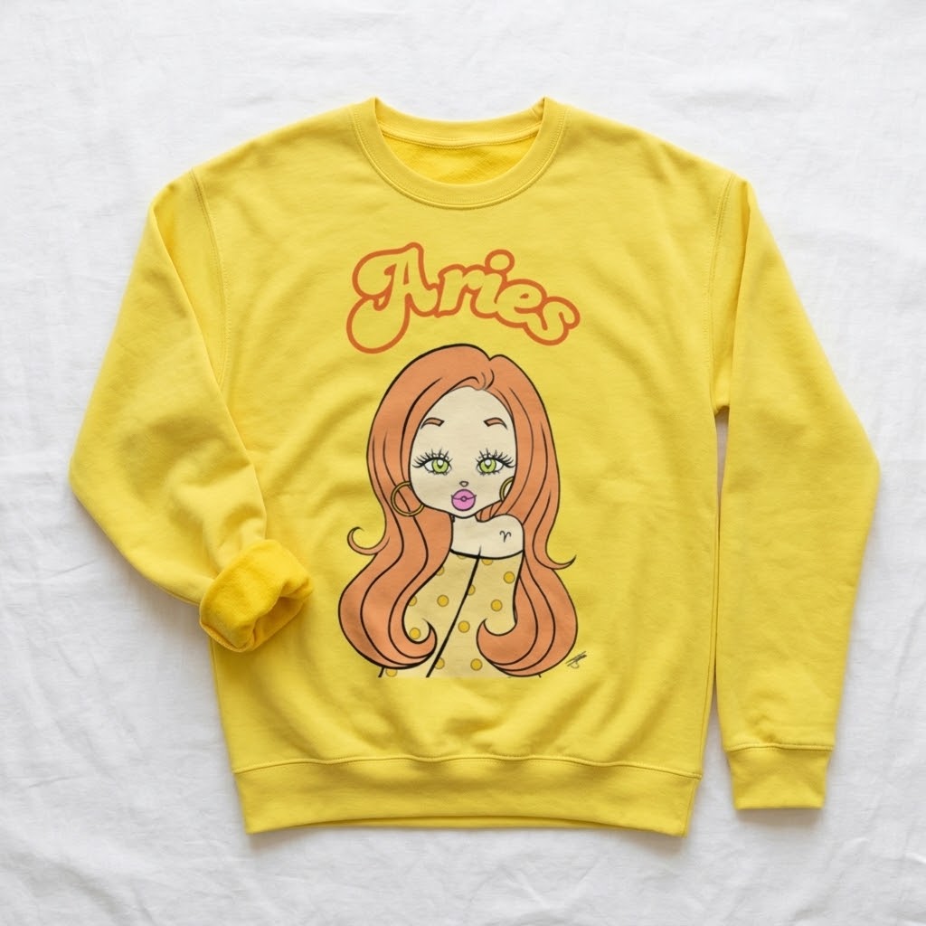 A yellow crewneck sweatshirt featuring a cartoon illustration of a woman with long orange hair and the word 'Aries' in orange script above her, laid flat on a white fabric background.