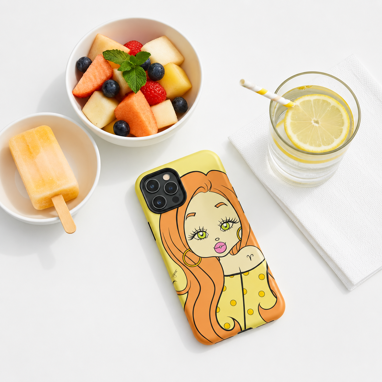 A yellow phone case with an Aries character design, styled in a flat-lay composition on a white surface alongside a bowl of fruit, a popsicle, and a glass of lemon water.