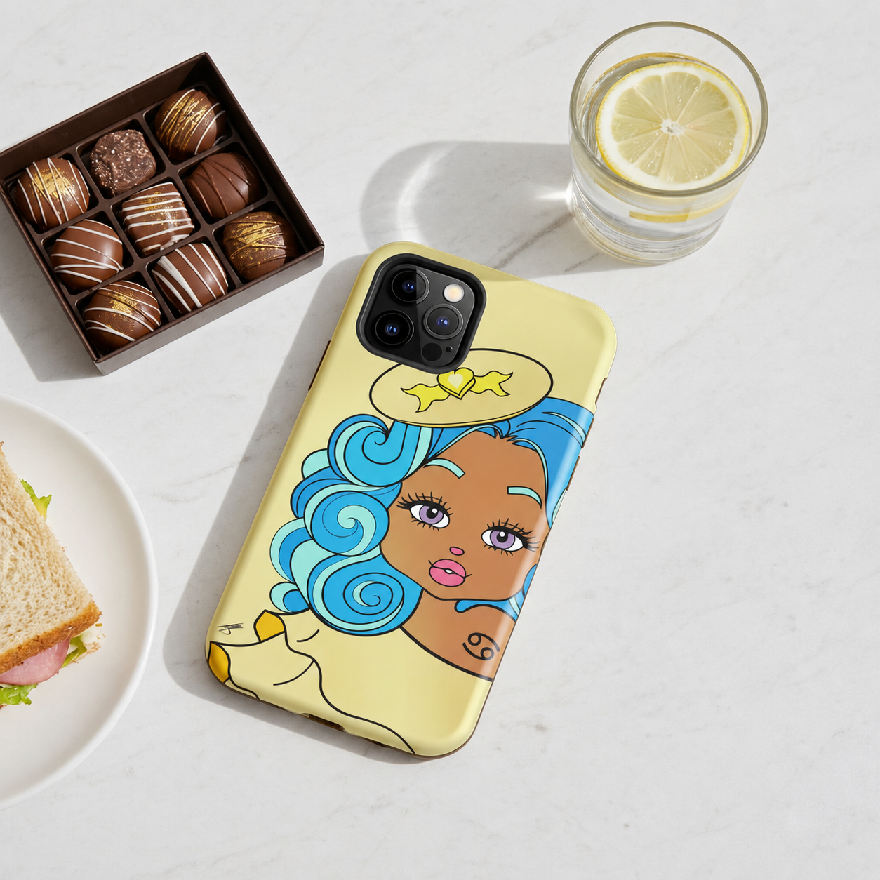 A light yellow phone case with a cancerian character design, shown on a white marble surface next to a chocolate box, a glass of lemon water, and a sandwich.