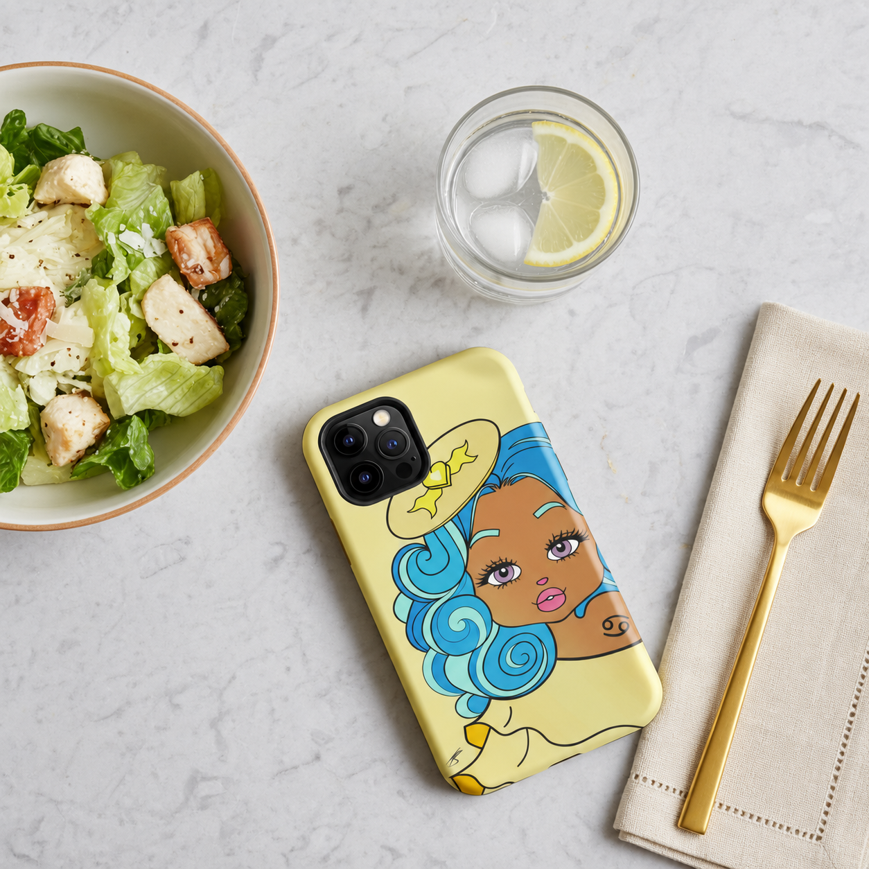 A yellow phone case featuring a cancerian character design placed on a light marble surface next to a salad bowl, a glass of water with a lemon slice, and a gold fork on a beige napkin.