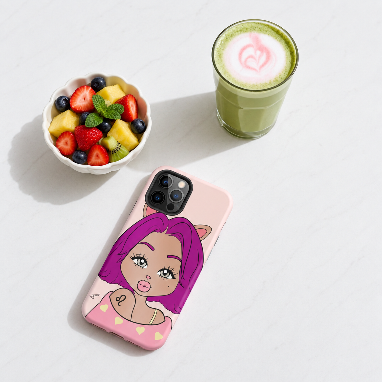 A light pink phone case with a leo character design, displayed on a white marble surface next to a bowl of fresh fruit and a green matcha drink.