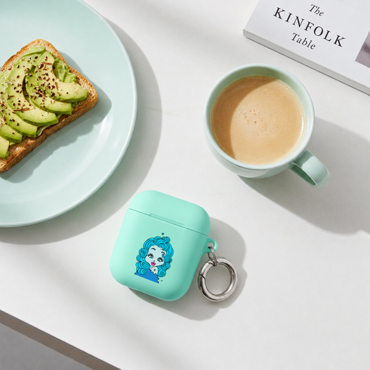 A mint green protective case for wireless earbuds with a Pisces character design and a silver key ring, styled on a white table next to a cup of coffee, avocado toast on a plate, and a book.