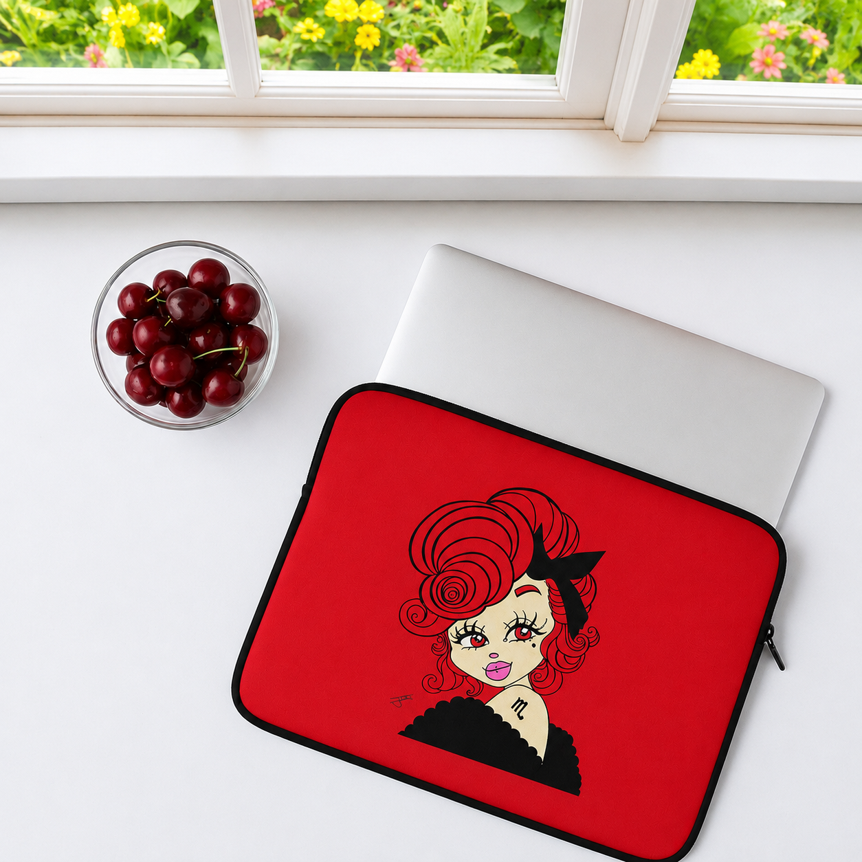 A red laptop sleeve with a leo character design, shown on a white desk next to a closed silver laptop and a small bowl of red cherries near a window.