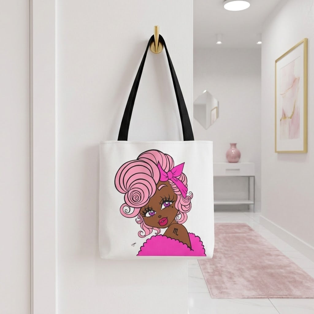 A white canvas tote bag with black shoulder straps hangs from a gold hook on a white wall. The bag features a colorful cartoon illustration of a woman with pink hair and a pink bow.