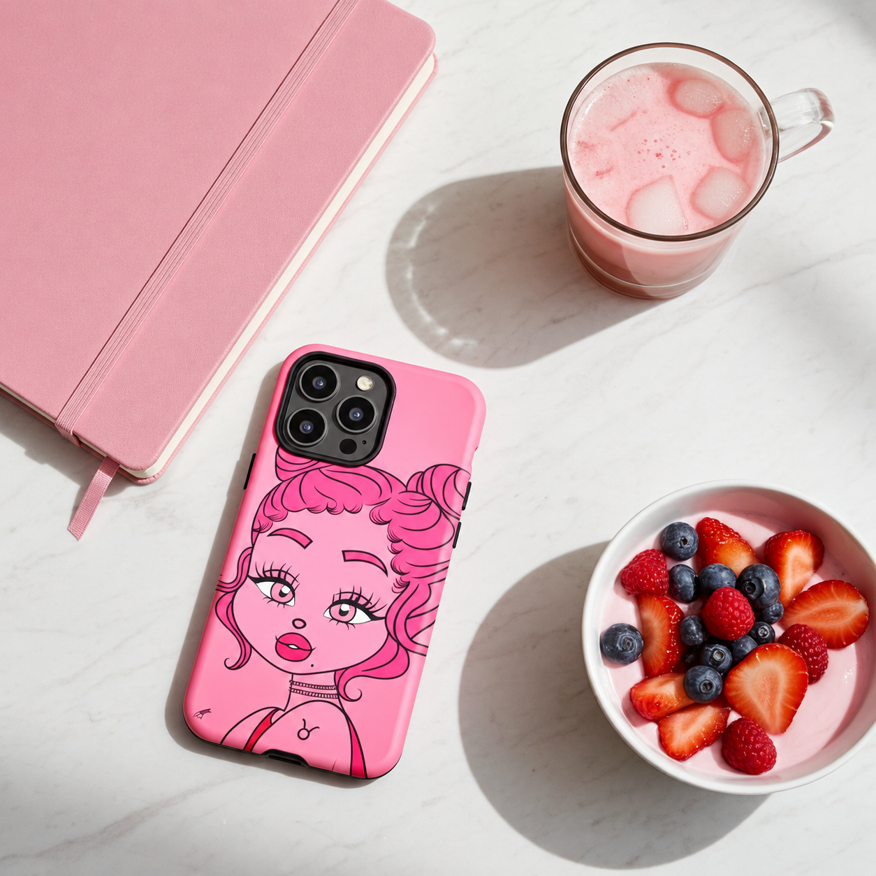 A pink phone case with a taurus character design on a white marble surface next to a pink notebook, a glass of pink drink, and a bowl of fruit yogurt.