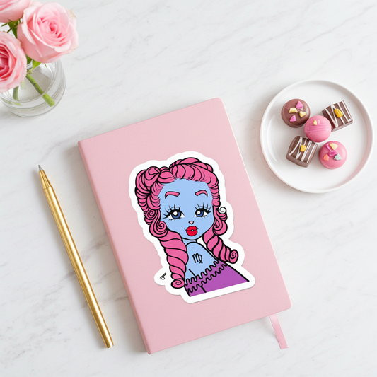 Pink notebook with a Virgo character sticker, gold pen, and plate of chocolates on a light surface.