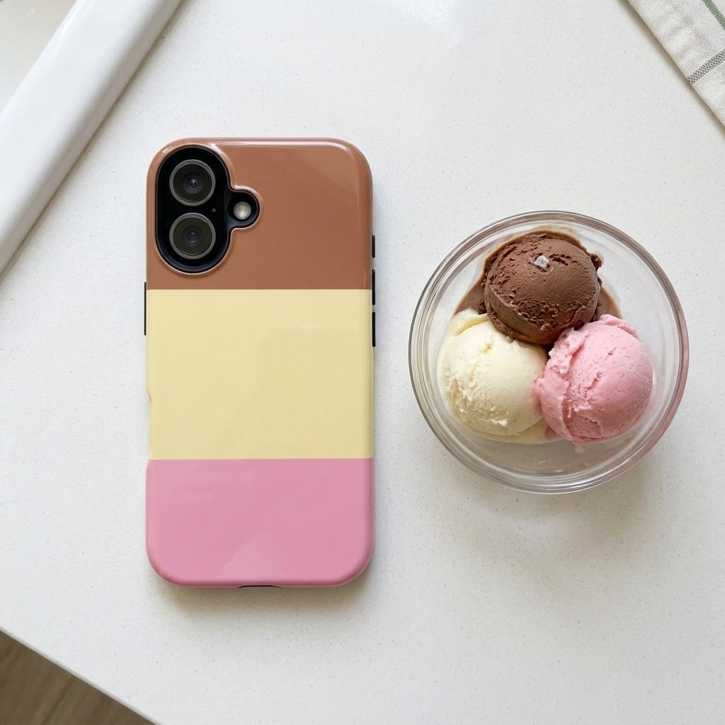 A phone case with a three-color block design in brown, pale yellow, and pink, shown next to a small glass bowl of ice cream on a white tabletop.