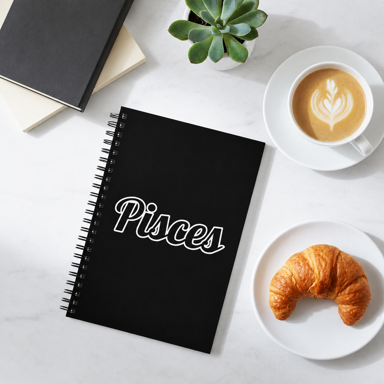 Black notebook with 'Pisces' on a white surface with a croissant and coffee.