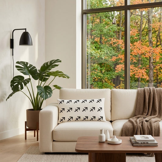 A cream-colored sofa in a modern living room setting, featuring a white throw pillow with a black Sagittarius pattern. The room includes a large window with a view of autumn trees, a potted monstera plant, a wooden coffee table, and a brown throw blanket.