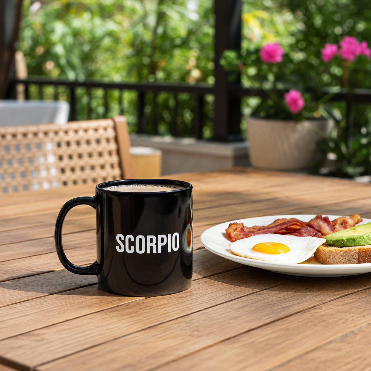 A black ceramic mug with the word 'SCORPIO' printed in white on the side sits on a wooden outdoor table next to a plate of breakfast food. The background shows a blurred patio setting with greenery and pink flowers.