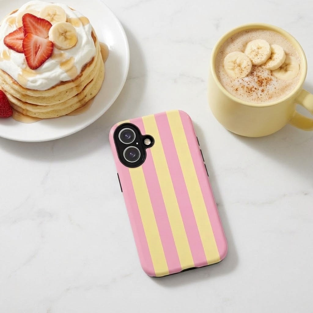 A phone case with vertical pink and pale yellow stripes is shown on a light marble surface, styled with a plate of pancakes topped with strawberries and whipped cream on the left and a yellow coffee mug with banana slices on the right.
