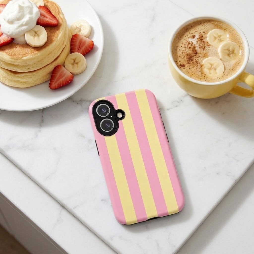 A phone case with vertical pink and pale yellow stripes, shown on a white marble surface next to a plate of pancakes and a cup of coffee with banana slices.