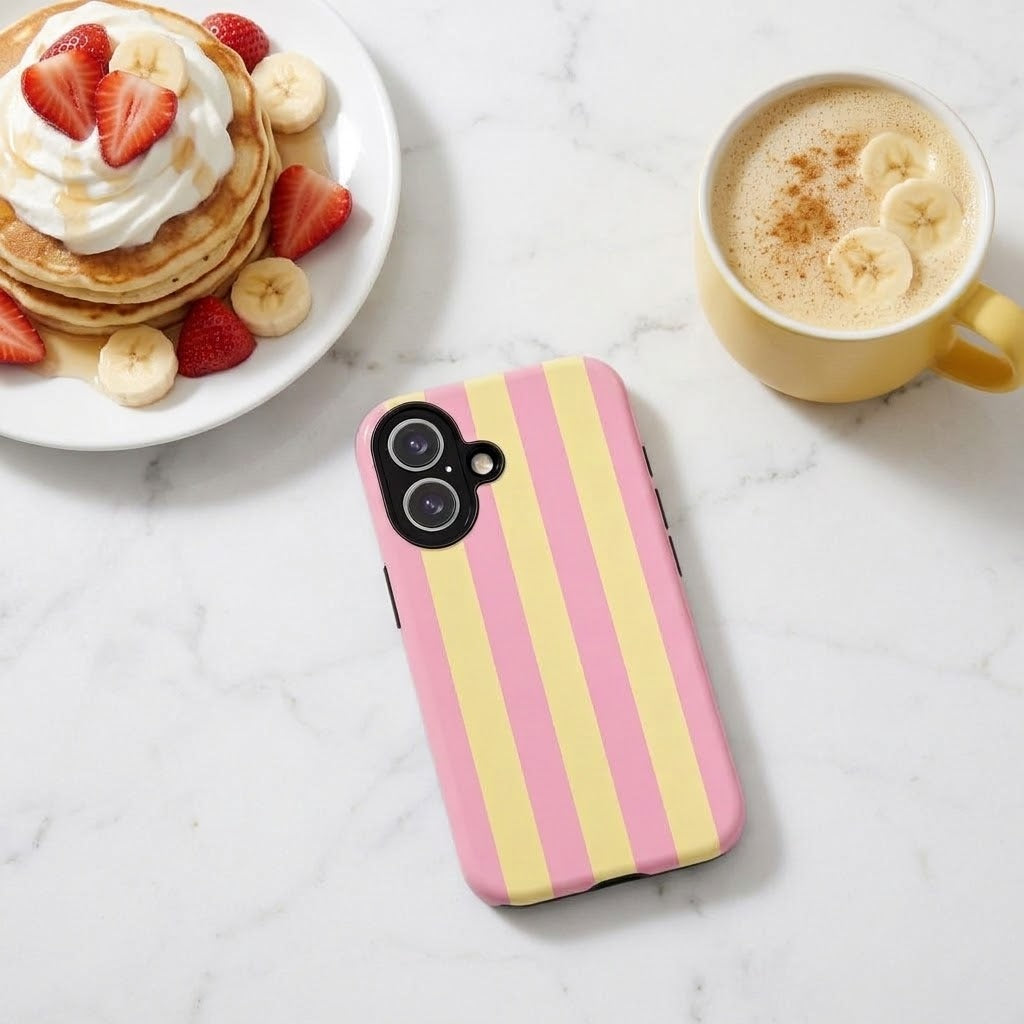 A pink and yellow striped phone case with a black camera cutout, displayed on a white marble surface next to a plate of pancakes with strawberries and bananas, and a yellow coffee mug.