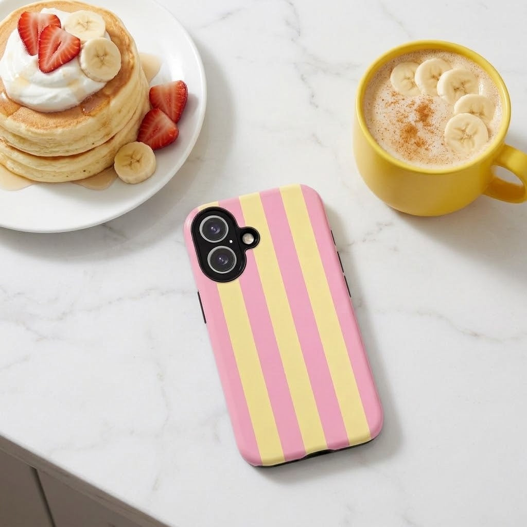 A pink and yellow vertically striped phone case with a camera cutout, displayed on a white marble surface next to a plate of pancakes and a yellow coffee mug.