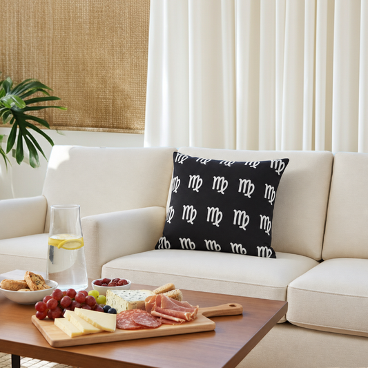 A black throw pillow with a repeating white Virgo pattern sits on a cream-colored sofa in a bright, modern living room setting with a charcuterie board and drinks on a wooden coffee table in the foreground.