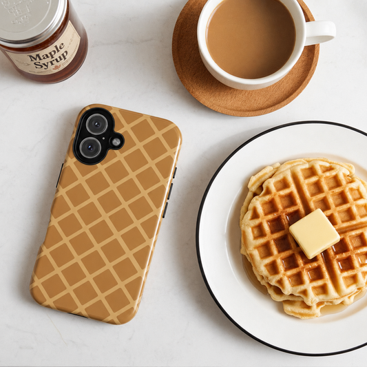 A tan and beige waffle-patterned phone case is displayed on a white marble tabletop alongside a cup of coffee, a plate of waffles with butter, and a jar of maple syrup.